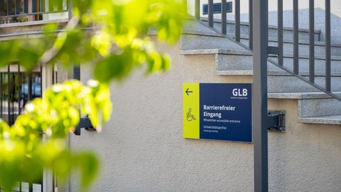 Das Foto zeigt ein Schild mit einem Pfeil und einem Rollstuhlfahrer zum barrierefreien Eingang zum Günther-Landgraf-Bau.