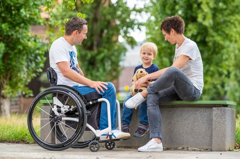 Foto zeigt einen Mann im Rollstuhl. Seine Frau und sein Kind sitzen neben ihm auf einer Bank.