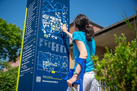 Foto einer Studentin mit Krücken, die versucht sich mit Hilfe einer Informationskarte auf dem TU Campus zu orientieren.