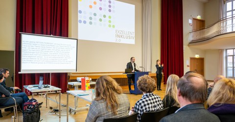 The photo shows a lecture in the Dülfersaal at TU Dresden from the audience's perspective. The speaker and a sign language interpreter can be seen in the background, standing in front of a presentation entitled ‘Exzellent Inklusiv 2.0’. On the left of the