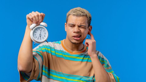 Ein junger Mann hält in der rechten Hand einen Wecker, während er sich mit der Linken ein Ohr zuhält. Sein Gesichtsausdruck ist verwirrt, er trägt ein gestreiftes T-Shirt. 