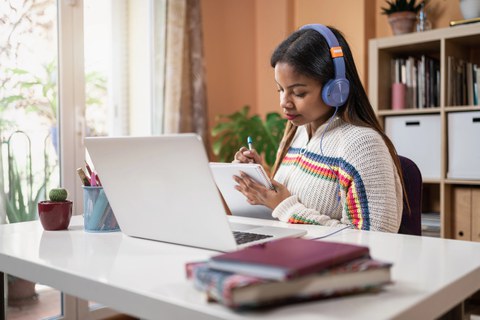 Auf dem Foto ist eine Person an einem Schreibtisch zu sehen. Sie hat Kopfhörer auf, vor ihr steht ein aufgeklappter Laptop und in ihrer Hand ist ein Block auf den sie schreibt. Am Rand des Tisches liegen Bücher. 