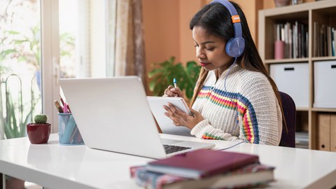 The photo shows a person sitting at a desk. They are  wearing headphones, have a laptop open in front of them, and are holding a notepad in their hand, on which they are writing. There are books lying on the edge of the desk. 