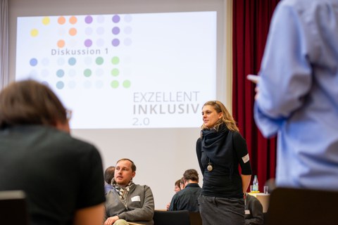 Eine Person steht in einer Diskussionsrunde und spricht mit den Teilnehmenden eines Workshops. Im Hintergrund ist eine Präsentationsfolie mit der Aufschrift ‚Diskussion 1‘ und ‚Exzellent Inklusiv 2.0‘ zu sehen. Mehrere Personen sitzen oder stehen im Raum 