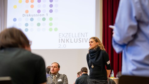 Eine Person steht in einer Diskussionsrunde und spricht mit den Teilnehmenden eines Workshops. Im Hintergrund ist eine Präsentationsfolie mit der Aufschrift ‚Diskussion 1‘ und ‚Exzellent Inklusiv 2.0‘ zu sehen. Mehrere Personen sitzen oder stehen im Raum 