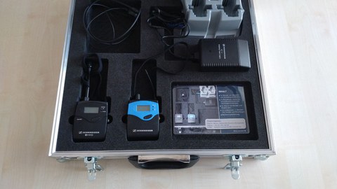 Photo shows the opened aluminum case placed on the floor. The case contains an FM system, a set consisting of a Sennheiser EW 300 G2 transmitter, a mobile receiver and a charging station.