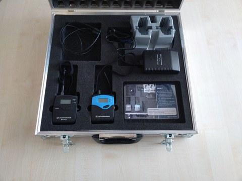 Photo shows the opened aluminum case placed on the floor. The case contains an FM system, a set consisting of a Sennheiser EW 300 G2 transmitter, a mobile receiver and a charging station.