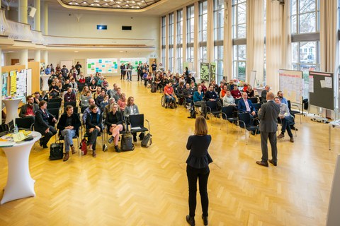 Das Foto zeigt einen Vortrag im Dülfersaal der TU Dresden. Während die eine Person in das Mikrofon spricht, gibt die Zweite dieses gesprochene Wort in Gebärdensprache wieder. Die Stuhlreihen im Hintergrund sind gut gefüllt.