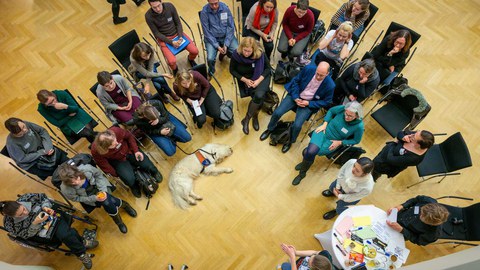 Blick auf eine laufende Diskussionsrunde von oben. Mittig liegt ein Assistenzhund. 