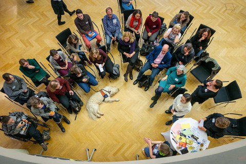 Blick auf eine laufende Diskussionsrunde von oben. Mittig liegt ein Assistenzhund. 