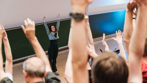 Beim Science Slam 2025 in Dresden steht eine Vortragende vorne im Hörsaal und animiert das Publikum, die Arme zu heben. Viele Zuhörer machen mit, wodurch eine lebendige, interaktive Stimmung entsteht.