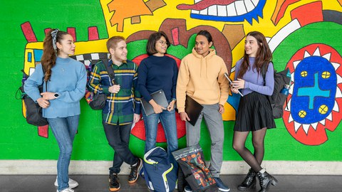 Foto von fünf Studierenden an der TUD, die sich vor einer großen bunten Wand unterhalten.