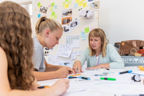 Auf dem Foto sind 3 Frauen zus ehen die an einem Schreibtisch sitzen und über eine Entwurf diskustieren. Im Hintergrund ist eine große Pinwand an der verschiedene Entwürfe hängen.