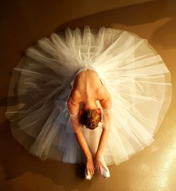 Das Foto zeigt eine Aufnahme einer Ballettänzerin von oben, die mit weit aufgefächertem Balletrock mit nach vorn gebeugten Oberkörper auf dem Boden sitzt.