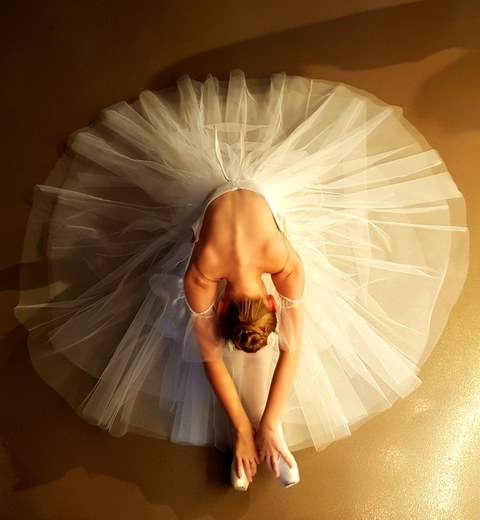Das Foto zeigt eine Aufnahme einer Ballettänzerin von oben, die mit weit aufgefächertem Balletrock mit nach vorn gebeugten Oberkörper auf dem Boden sitzt.