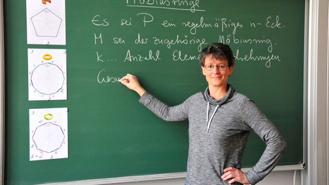 Das Foto zeigt eine Frau (Dr. Antje Noack) vor einer grünen Schultafel, an die sie einen Satz über Möbiusringe geschrieben hat.