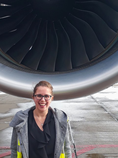 Das Bild zeigt eine Frau vor einem riesigen Flugzeugtriebwerk.