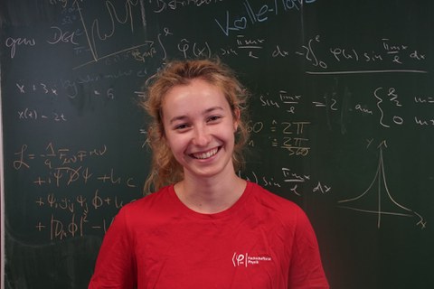 Das Bild zeigt eine junge blonde Frau vor einer grünen  Tafel an der viele verschiedene mathematische Gleichungen und Formeln geschrieben sind.