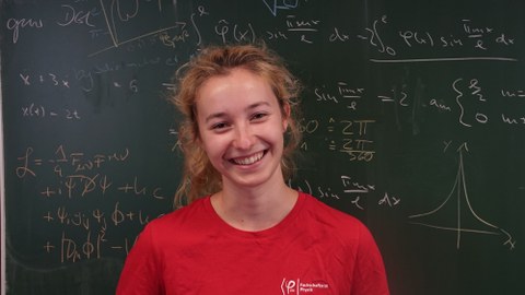 Das Bild zeigt eine junge blonde Frau vor einer grünen  Tafel an der viele verschiedene mathematische Gleichungen und Formeln geschrieben sind.