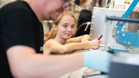 Das Foto zeigt eine junge blonde Frau die an einer Maschine hockt und etwas aufschreibt.