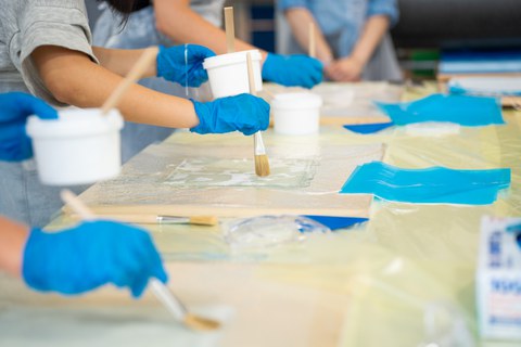 Das Foto zeigt Hände von Personen mit blauen Handschuhen, die mit einem Pinsel etwas auftragen.