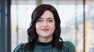 Bild einer Frau, die an einem Geländer lehnt, dunkle offene Haare hat und ein grünes Oberteil trägt. Sie lächelt.