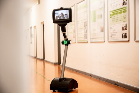  Das Bild zeigt einen Telepräsenzroboter, der in einem Korridor steht. Der Roboter hat ein Display an der Spitze, auf dem das Gesicht einer Person zu sehen ist, die scheinbar gerade per Videoanruf mit dem Roboter verbunden ist. Der Roboter besteht aus einem stabilen Standfuß mit Rollen am Boden, einem schmalen Mittelteil und einem beweglichen Kopf, der das Display hält. An einer Stelle am Mittelteil des Roboters befindet sich ein leuchtendes grünes Licht.  Der Korridor, in dem der Roboter steht, ist mit hellem Bodenbelag versehen, und an den Wänden hängen Informationsplakate oder Poster, die sich in Rahmen befinden. Die Wände des Korridors sind in einer hellen Farbe gestrichen. Die Türrahmen sind dunkel gehalten und verleihen dem Raum einen klaren, professionellen Eindruck. Der Roboter ist allein im Bild und scheint sich in einer ruhigen Umgebung zu befinden, möglicherweise in einem Bürogebäude oder einer Bildungseinrichtung.