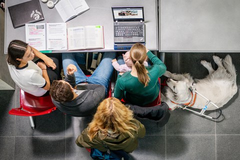 The photo, taken from above, shows three students sitting side by side on red chairs at a gray desk. A fourth student is standing behind them, also facing the desk. The student on the right has a baby on her lap and guide dog is sitting at her feet.