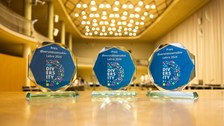 Three glass awards labeled "Preis diversitätssensible Lehre 2024" are displayed on a wooden table in a well-lit hall.