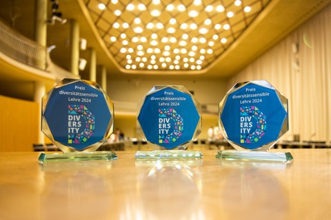 Three glass awards labeled "Preis diversitätssensible Lehre 2024" are displayed on a wooden table in a well-lit hall.
