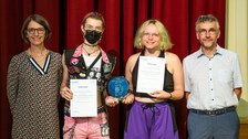 Four people stand together and two people hold an award and two certificates in their hands
