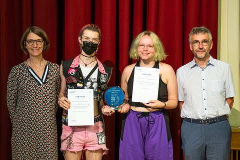 Four people stand together and two people hold an award and two certificates in their hands