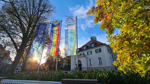 Drei Flaggen vor einem hellen hisorischen Gebäude. Die linke Flagge zeigt das Logo der TU Dresden, die Mittlere zeigt die Pride-Flag und die Drrtte Flagge verweist auf den Kooperationspartner Dresden CONCEPT e.V.