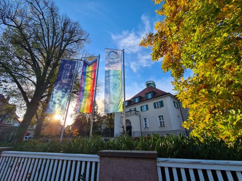 Drei Flaggen vor einem hellen hisorischen Gebäude. Die linke Flagge zeigt das Logo der TU Dresden, die Mittlere zeigt die Pride-Flag und die Drrtte Flagge verweist auf den Kooperationspartner Dresden CONCEPT e.V.