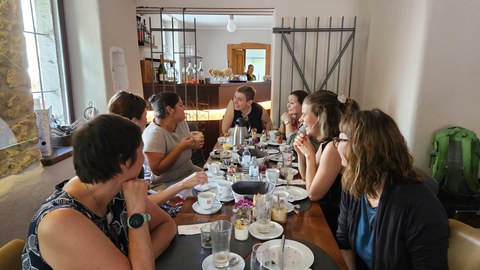 Mehrere Frauen sitzen an einer langen Tafel in einem gemütlichen Restaurant und unterhalten sich beim Frühstück. Auf dem Tisch stehen Teller, Tassen, Gläser und verschiedene Speisen.  