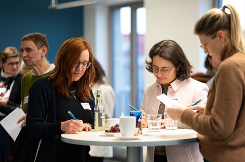 Mehrere Frauen stehen an einem Stehtisch beim Neujahrsempfang des Women@DDc Netzwerks, schreiben Notizen und tauschen sich in entspannter Atmosphäre bei Getränken aus.