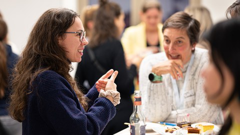 Zwei Frauen sitzen an einem Tisch beim Neujahrsempfang des Women@DDc Netzwerks, unterhalten sich lebhaft und gestikulieren, während auf dem Tisch Getränke und Kuchen stehen.