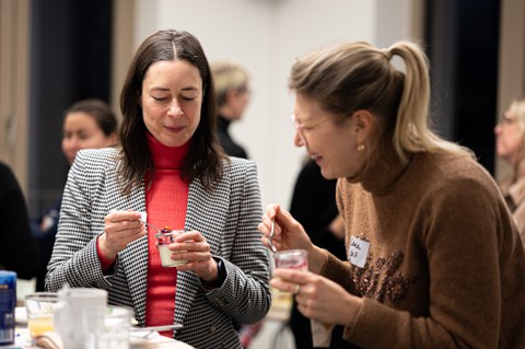 Zwei Frauen stehen an einem Stehtisch, lachen zusammen und löffeln ein Dessert aus einem Glas.
