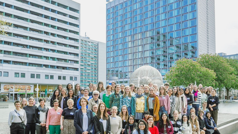 Große Gruppe draußen vor modernen Gebäuden für ein Gruppenfoto versammelt, viele Personen nebeneinander stehend.