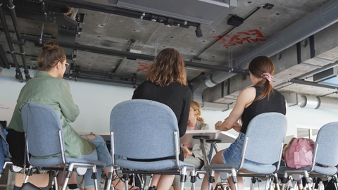 Gruppe sitzt in modernem Raum auf Stühlen um einen Tisch, Rücken zur Kamera, konzentrierte Gesprächssituation.