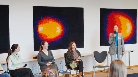 Podiumsrunde in hellem Raum, vier Personen sitzen, eine Person steht mit Mikrofon und spricht. Bilder mit farbigen Kreisen hängen an der Wand.