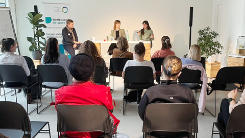 Publikum sitzt vor drei Personen auf kleiner Bühne, eine spricht, zwei hören zu; heller Raum mit Technik über dem Podium.