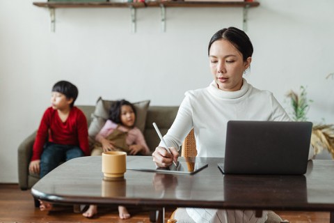 Frau sitzt an einem Schreibtisch und im Hintergrund sitzen zwei Kinder auf einer Couch