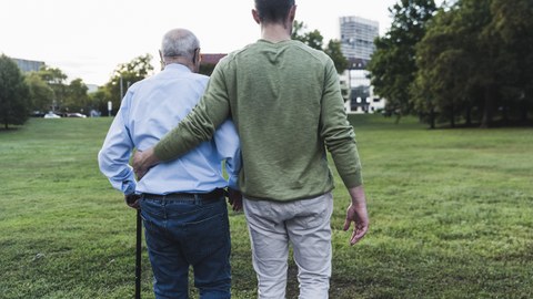 Picture of a grandfather with a cane being supported by his grandson and walking with him across a meadow.