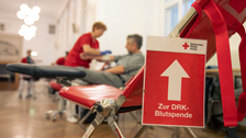 An einem Stuhl hängt ein rotes Schild des deutschen Roten Kreuzes mit einem weißen Pfeil. Darunter ist weißer Schrift zu lesen "Zur Blutspende". Im Hintergrund erkennt man eine Person, die der anderen Blut abnimmt.