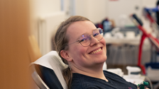 Eine Person sitzt auf einer Liege. Ihr wird Blut abgenommen. Sie lacht strahlend in die Kamera