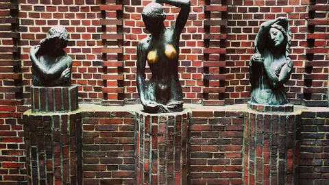 The image shows three bronze female busts mounted on brick pedestals against a red brick wall. The central statue stands out with its polished breasts, suggesting frequent touch by visitors. 