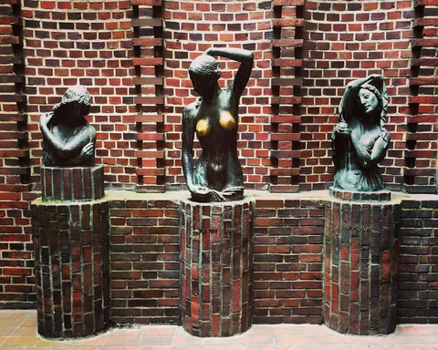 Auf dem Bild sind drei bronzene Frauenbüsten zu sehen, die auf gemauerten Ziegelpfeilern vor einer rotbraunen Backsteinwand stehen. Die mittlere Statue fällt durch ihre glänzend polierten Brüste auf, die auf häufige Berührungen hindeuten. 