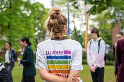 Eine Person trägt einen hohen Dutt. Sie trägt ein weißes T-Shirt. Man sieht sie nur von hinten. Auf dem shirt steht in Regenbogenfarben Lovers unite