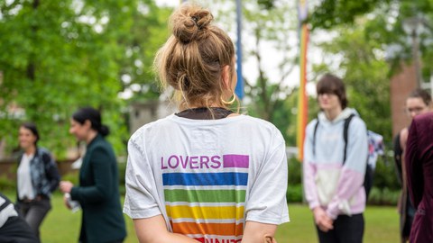 Eine Person trägt einen hohen Dutt. Sie trägt ein weißes T-Shirt. Man sieht sie nur von hinten. Auf dem shirt steht in Regenbogenfarben Lovers unite